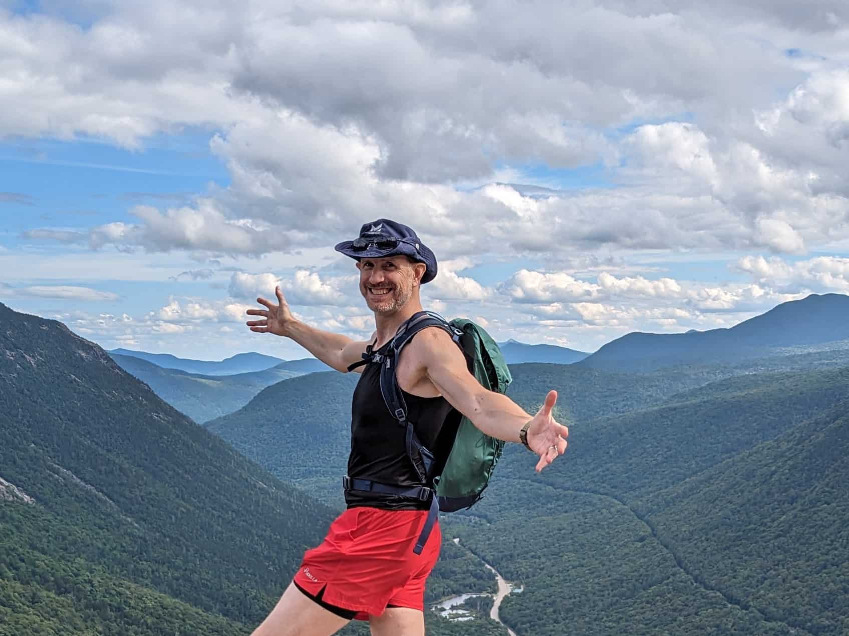 Mickey Mullin standing atop Mount Willard in NH, overlooking the valley below, arms spread wide.