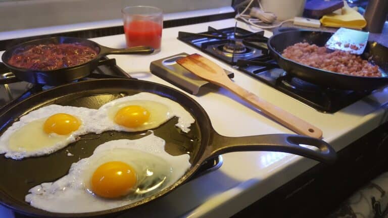 Eggs frying in a cast iron skillet beside a fruit compote small cast iron saucepan and corned beef hash in another skillet.