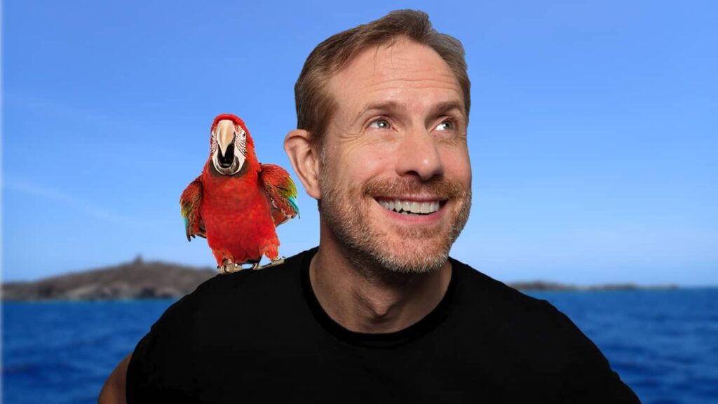 Mickey in black T-shirt against a blue Caribbean sea with an island in the background. There's a red green wing macaw on his shoulder